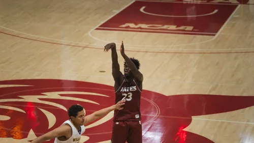A Bates College basketball player in a maroon jersey follows through on a jump shot as a defender reaches up beside him.