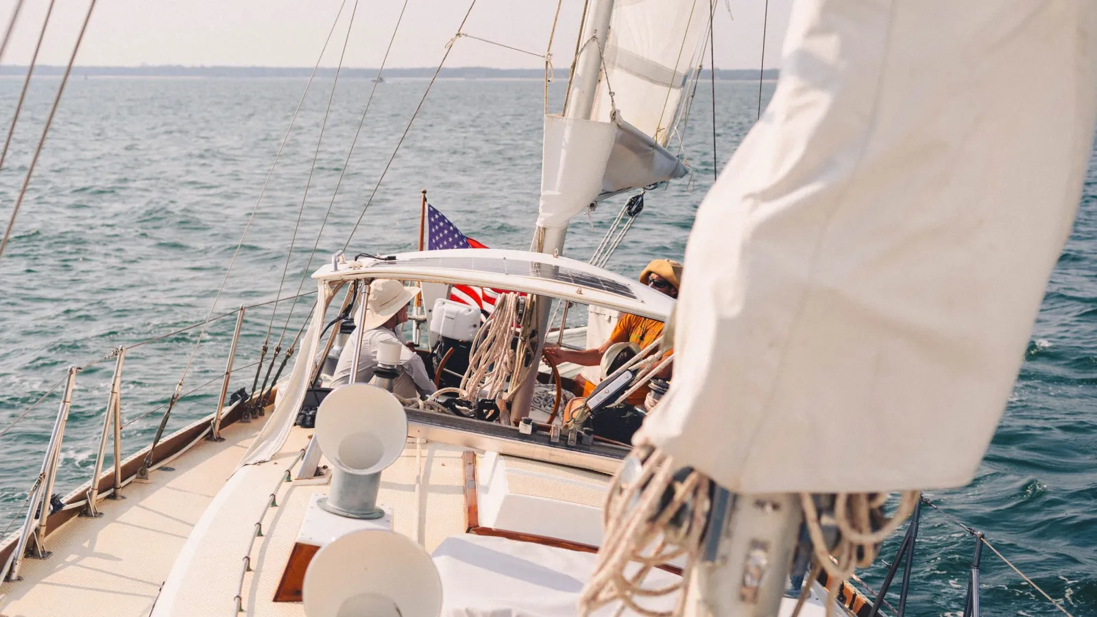 A person at the bow of a sailboat adjusting rigging near an American flag, open ocean and distant coastline behind.