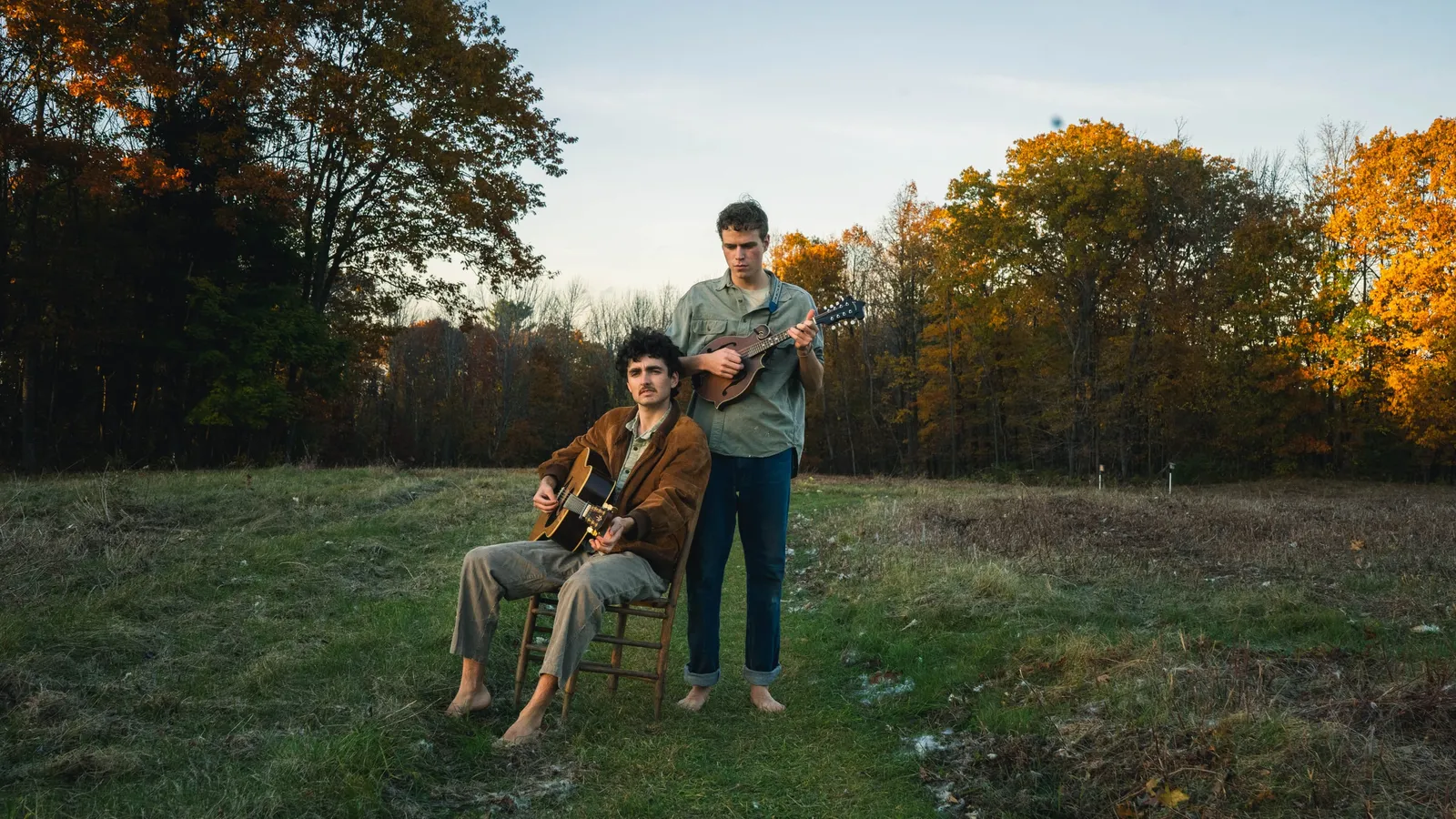Two young musicians in a field at golden hour, one seated barefoot playing acoustic guitar, the other standing playing mandolin.