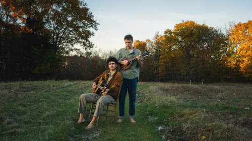 Two young musicians in a field at golden hour, one seated barefoot playing acoustic guitar, the other standing playing mandolin.