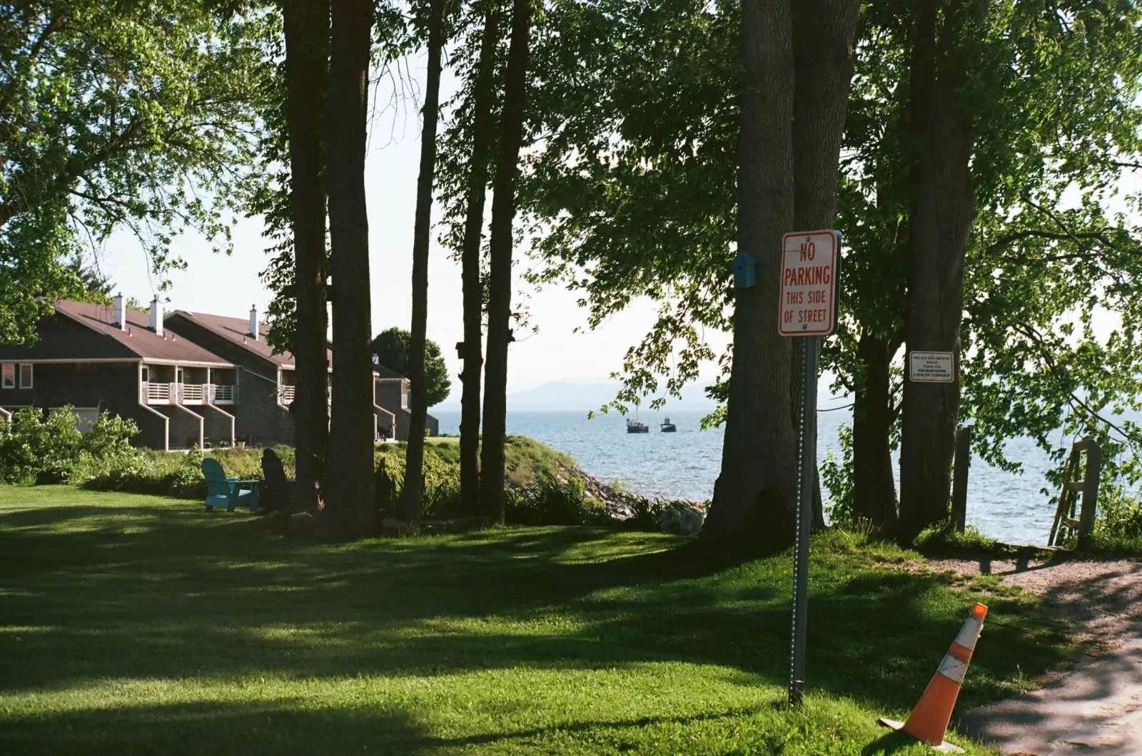 Film photo of a tree-lined lakeshore yard with a no-parking sign, cottages, and two small boats on the water.