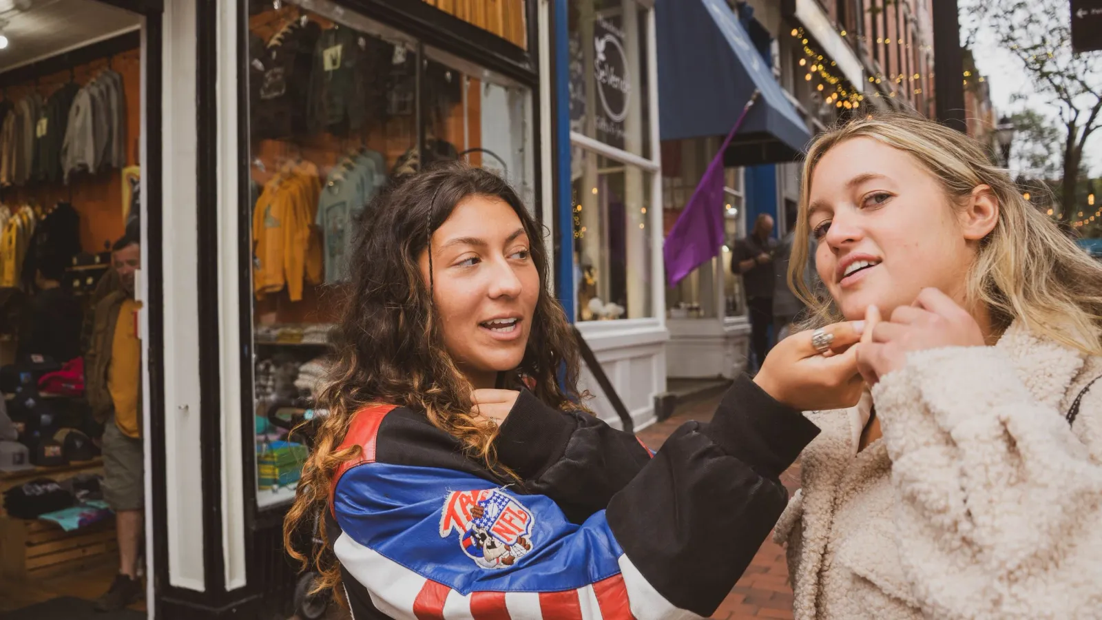 Two friends laughing on a sidewalk in front of storefronts, one adjusting the other's collar.