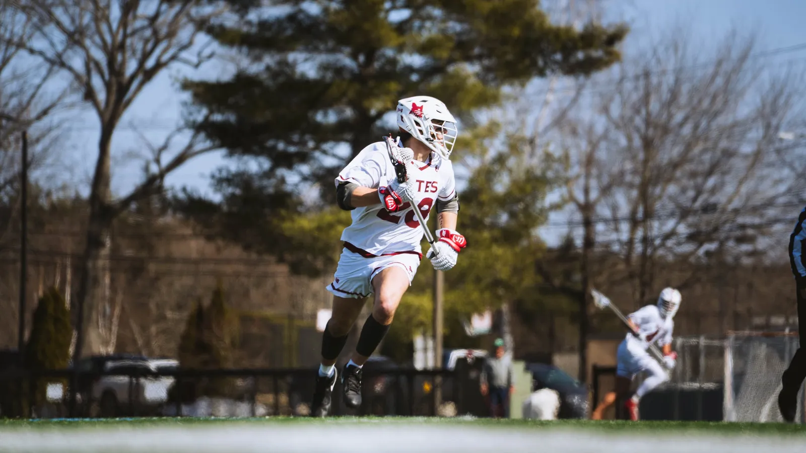 A Bates College men's lacrosse player in white sprints upfield carrying the ball in his stick, trees behind him.