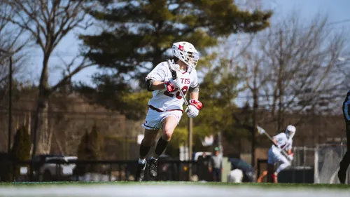 A Bates College men's lacrosse player in white sprints upfield carrying the ball in his stick, trees behind him.