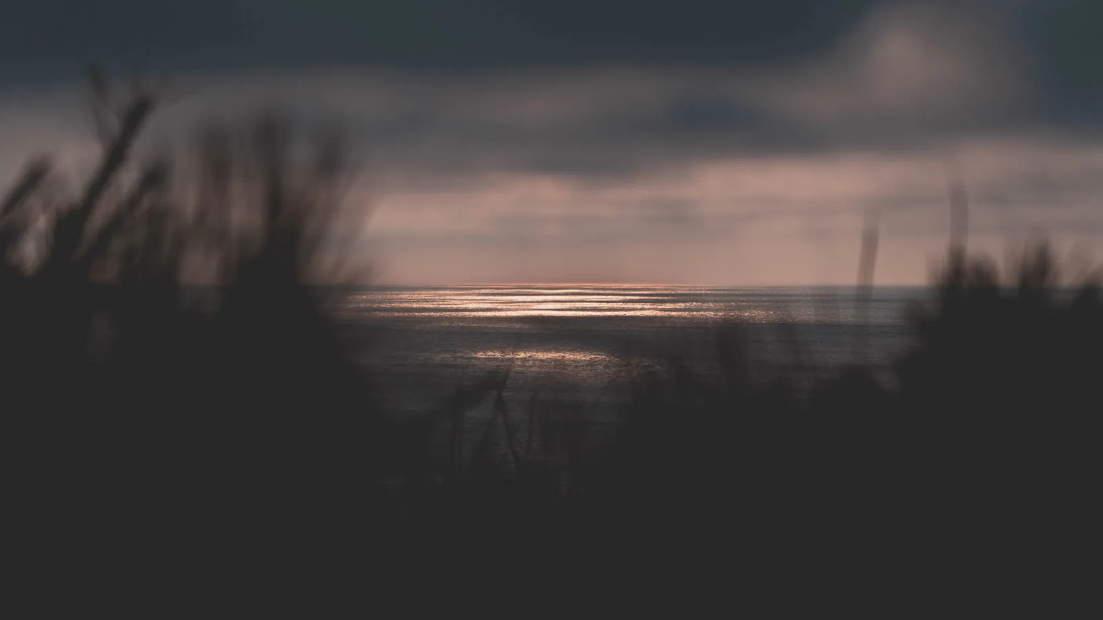 The sun setting into the ocean, its warm reflection stretching across calm water below a bank of clouds.