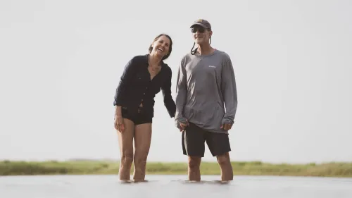 An older couple laughing together while standing ankle-deep in shallow water, holding hands.