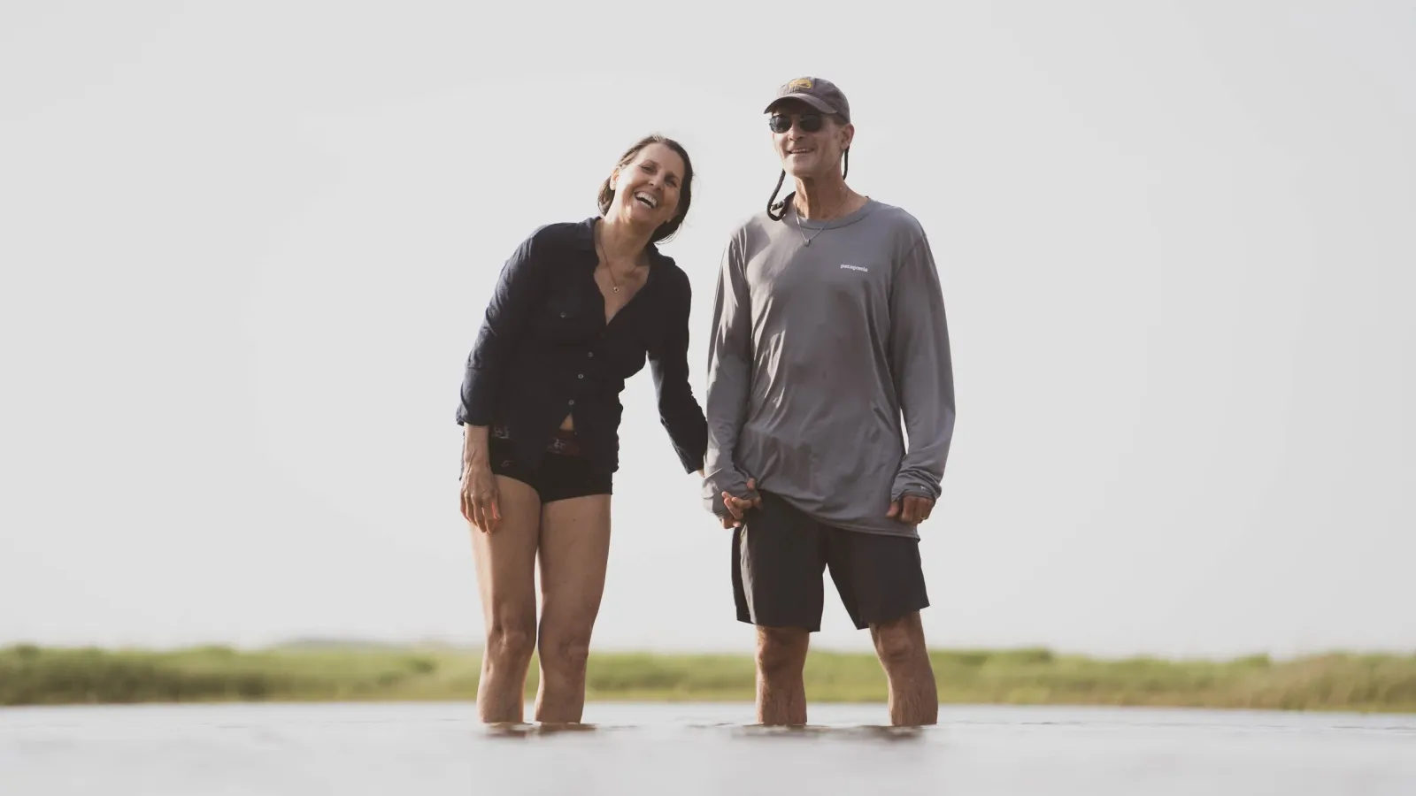 An older couple laughing together while standing ankle-deep in shallow water, holding hands.