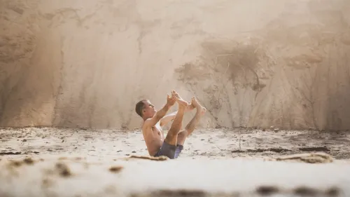 A shirtless man on a beach holding his feet overhead in a yoga pose, dust and light filtering past a sandy bluff behind him.