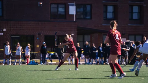 A Bates College field hockey player mid-swing on a hit, teammates and opponents nearby in front of a brick building.
