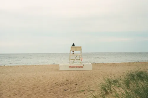 Film photo of a woman sitting atop a tall Nantucket lifeguard chair, facing the ocean.