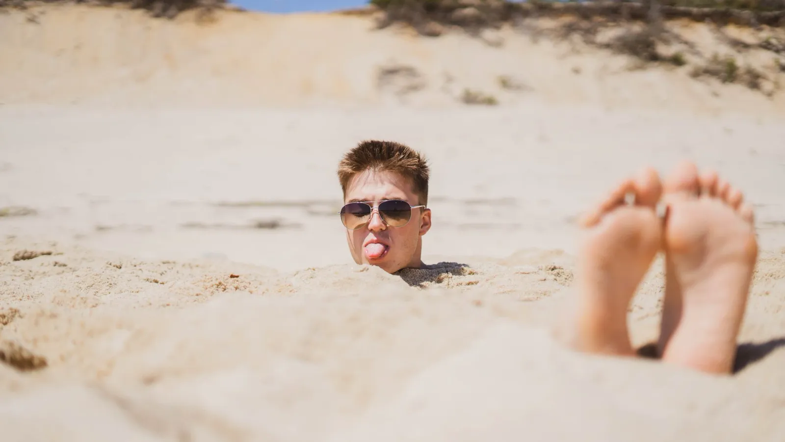A young man buried up to his neck in sand, sticking out his tongue, bare feet in the foreground.