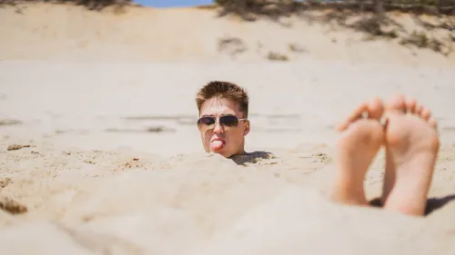 A young man buried up to his neck in sand, sticking out his tongue, bare feet in the foreground.