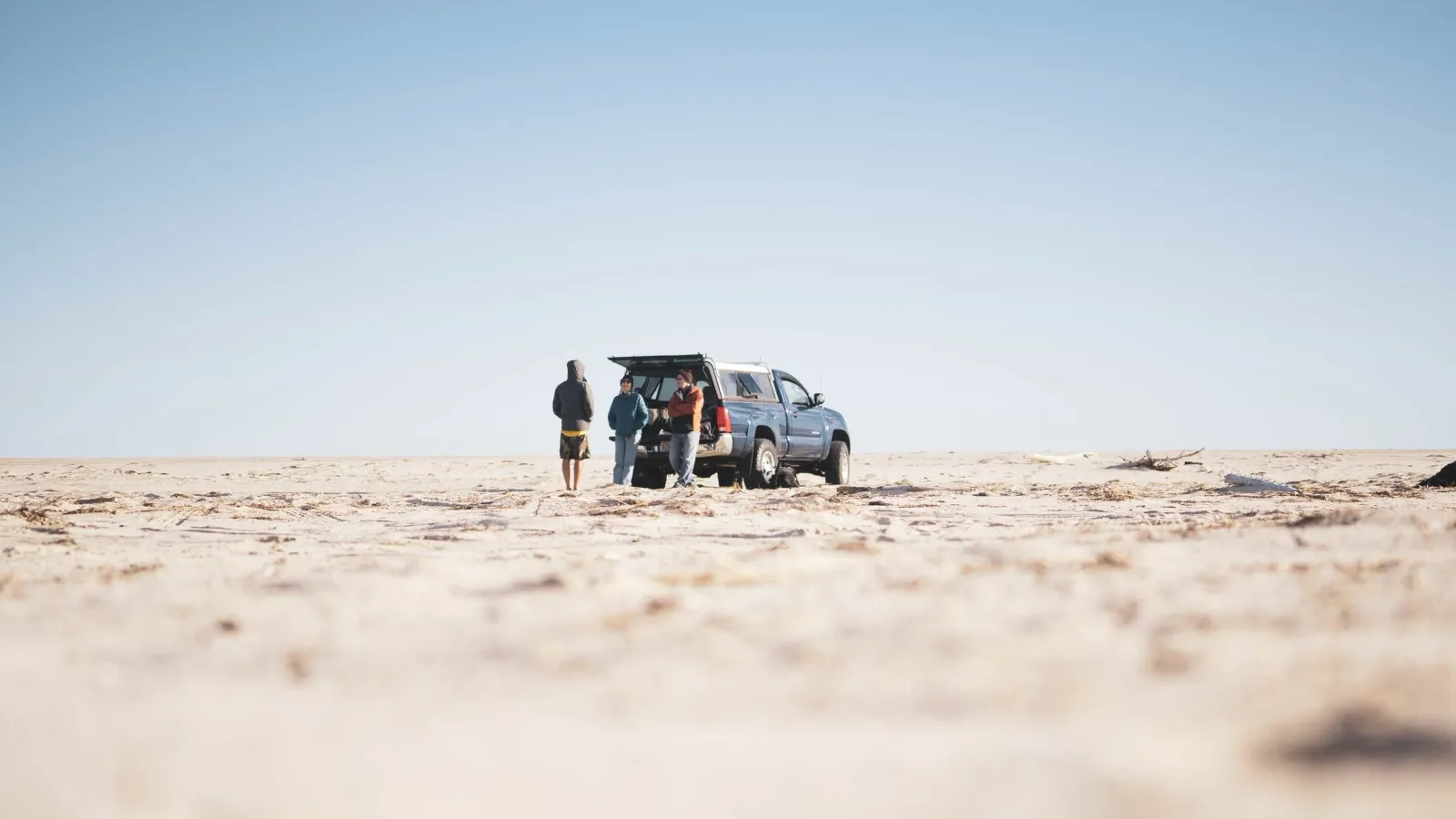 Wide desert landscape with three people and a pickup truck parked on open sand under a clear blue sky.