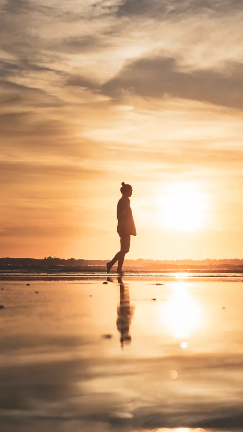 A woman's silhouette walking along a wet beach at sunset, her reflection mirrored in the sand.