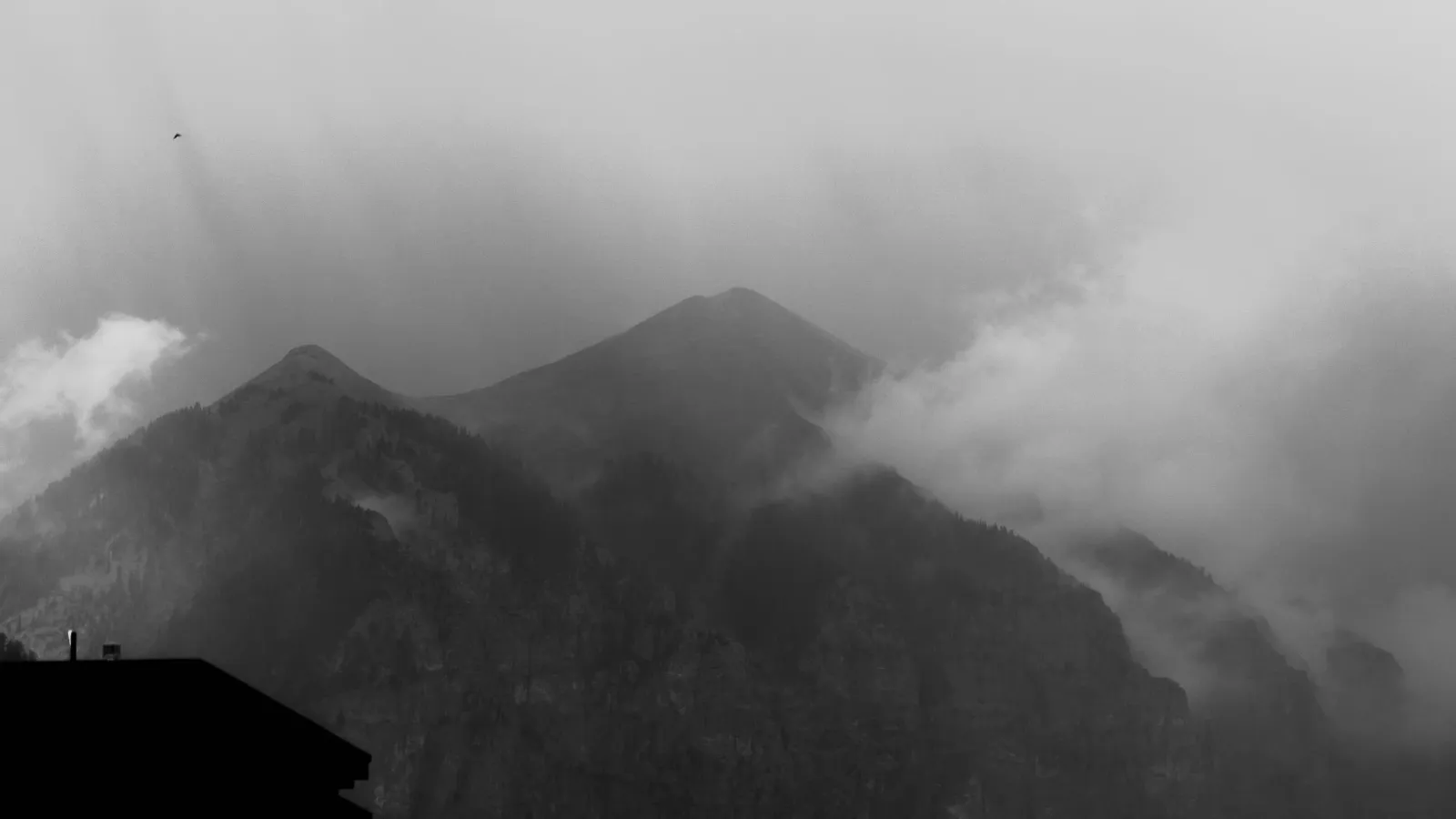 Black-and-white view of a mountain peak partly wrapped in cloud, a rooftop silhouette in the foreground.