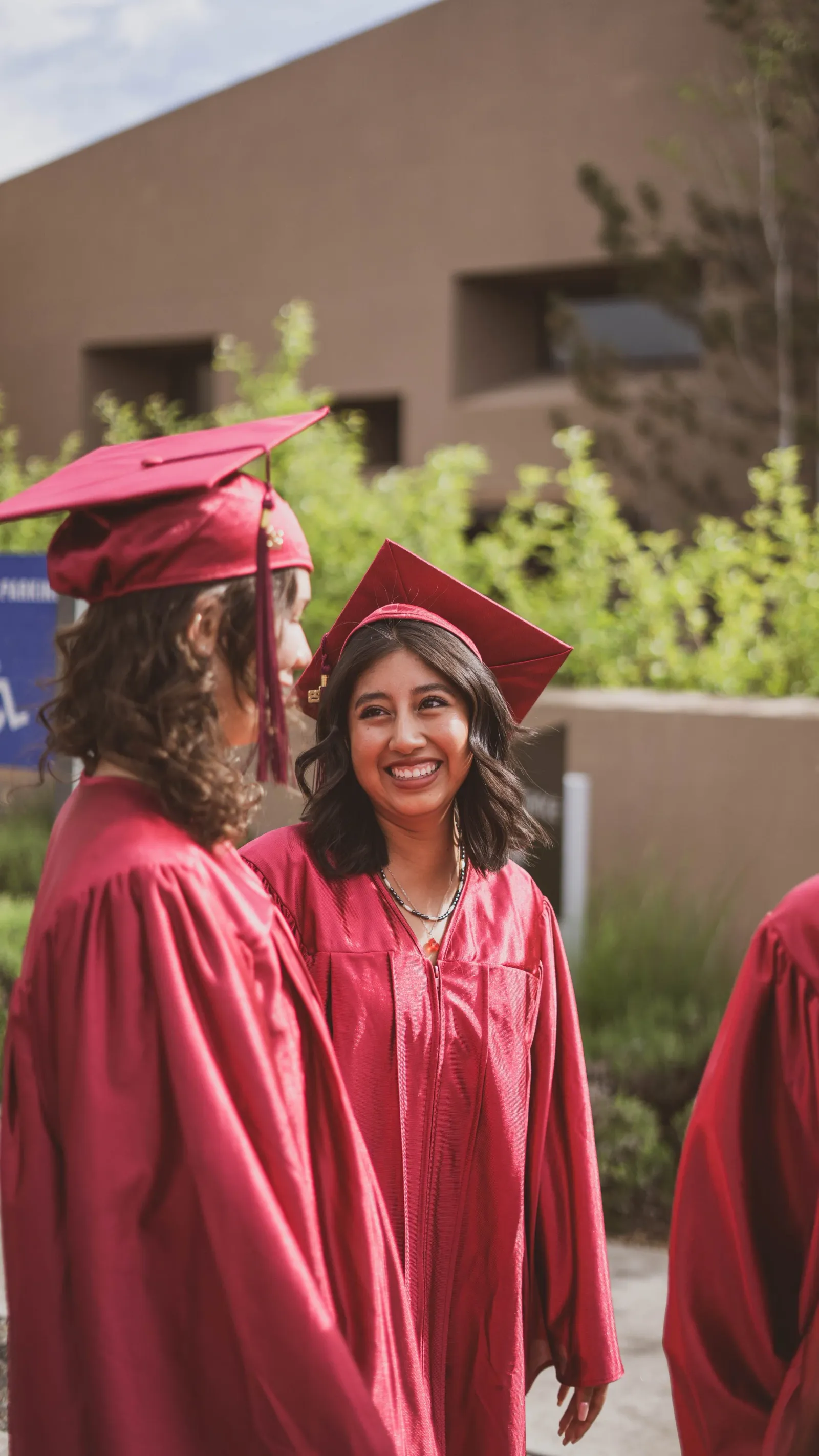 A young woman in a maroon cap and gown smiling at a graduation ceremony, another graduate beside her.