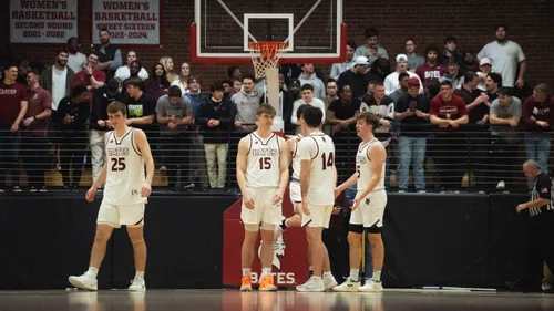 Four Bates College men's basketball players in white jerseys standing together on court in front of a packed crowd, championship banners overhead.