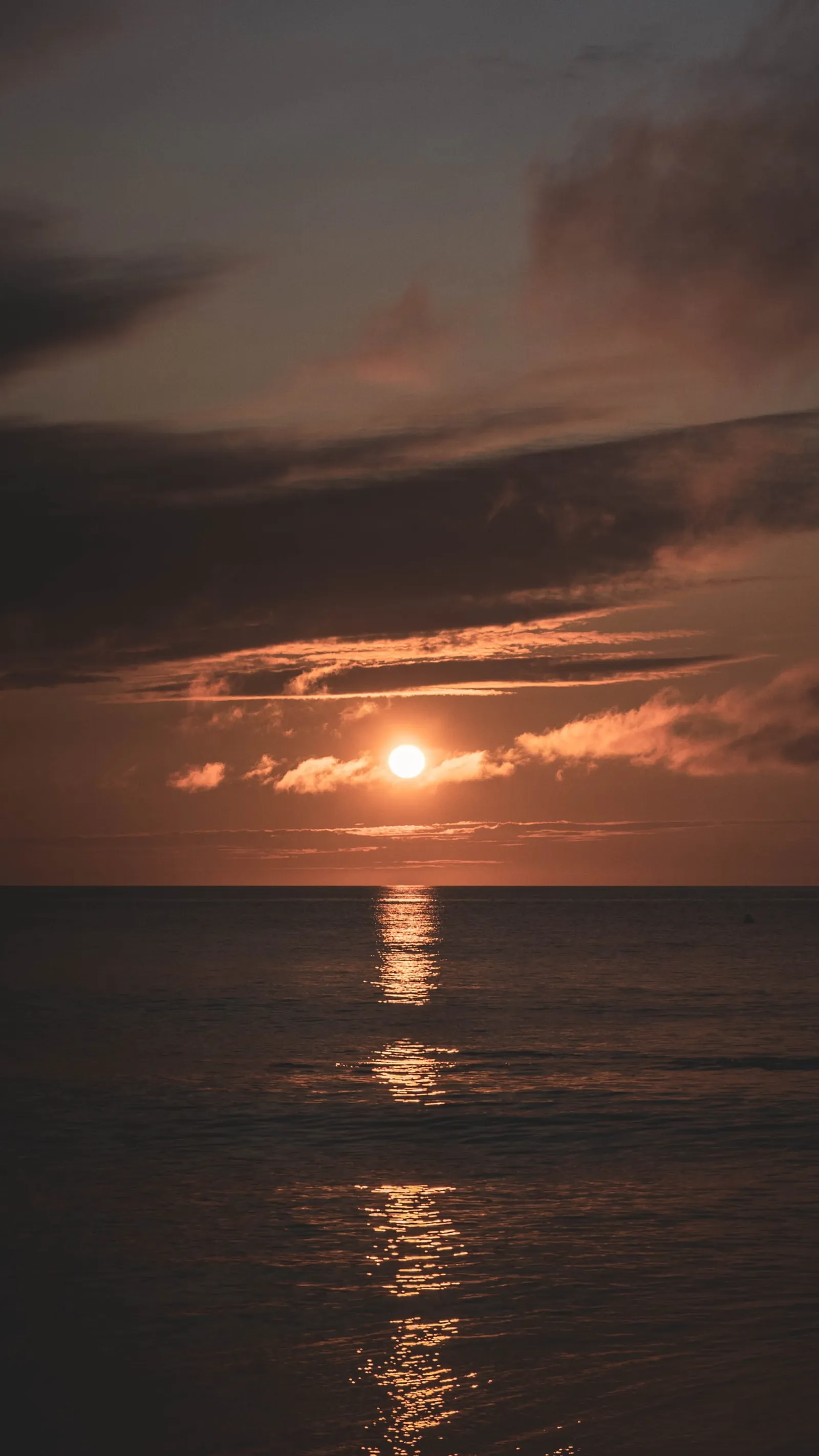 The sun setting behind a thin band of clouds over calm ocean water, its reflection stretched toward the shore.