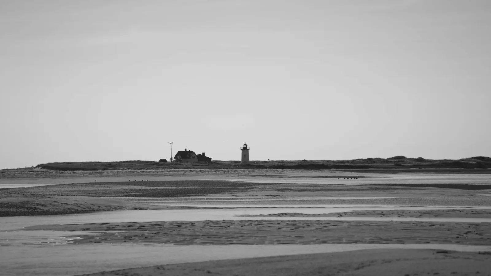 Black-and-white view of a lighthouse and keeper's house on a distant dune, seen across a wide tidal flat.