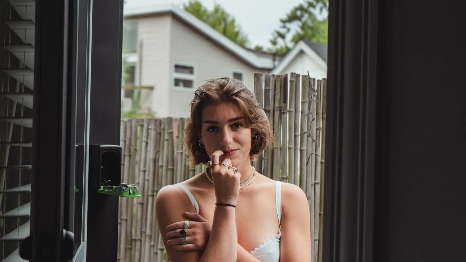 A young woman standing in a doorway with arms crossed, looking at the camera, a backyard bamboo fence and houses behind her.