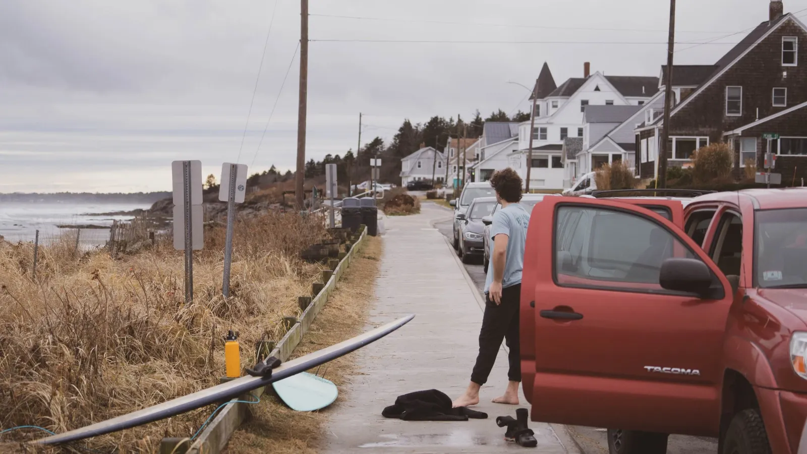 A man with a surfboard walking barefoot up a coastal street beside a red pickup truck.