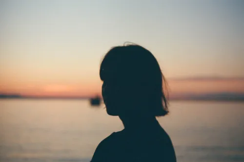 Film photo of a woman's silhouette against a pink and orange dusk sky over water, a small boat in the distance.