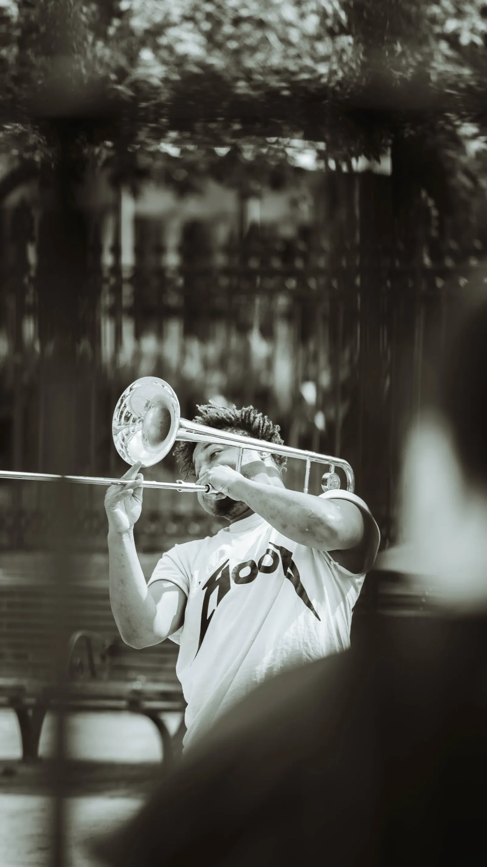 Black-and-white portrait of a street musician playing trombone through an iron fence.