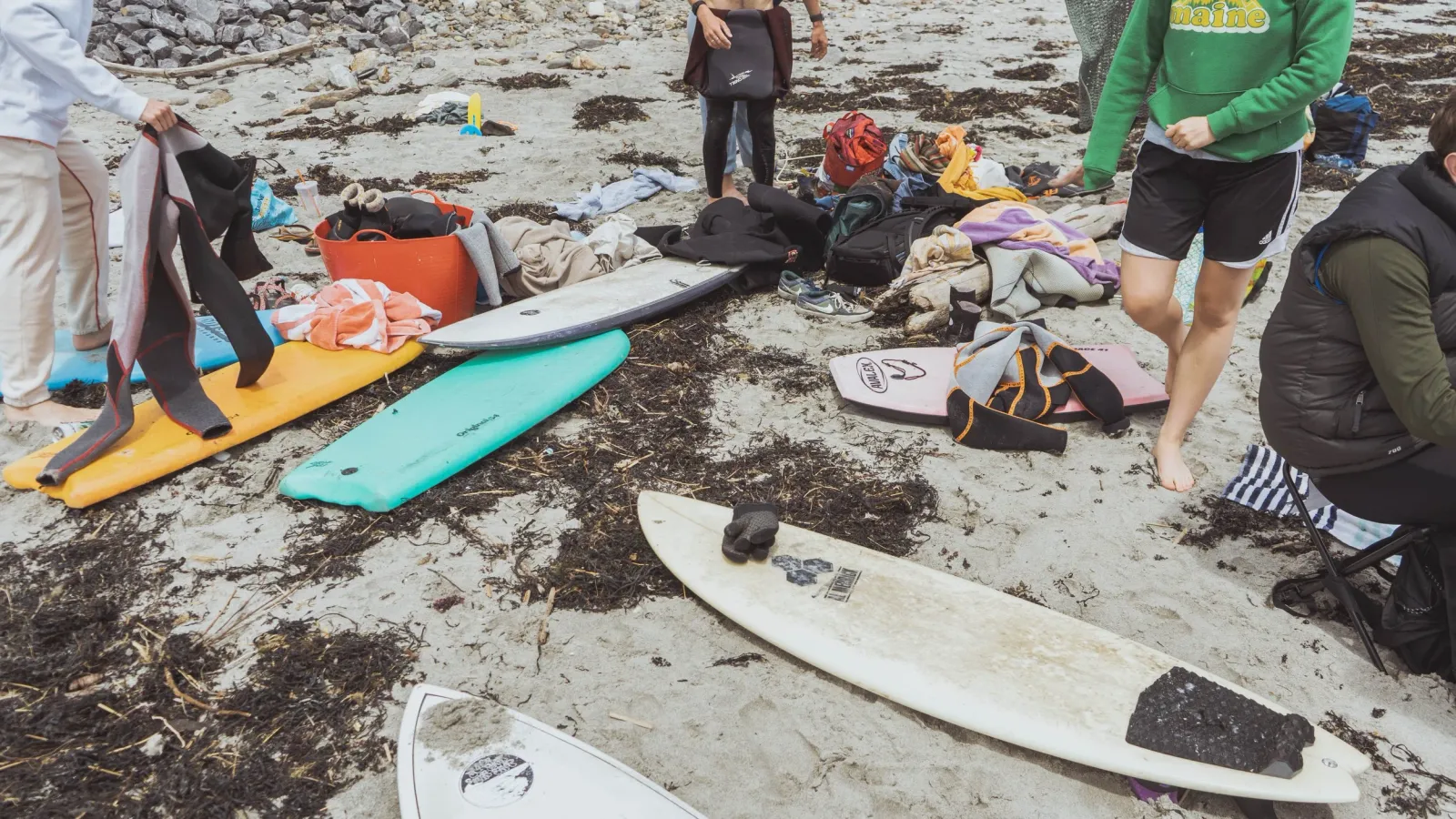 A group of people sorting through surfboards, wetsuits, and towels scattered on a rocky beach.