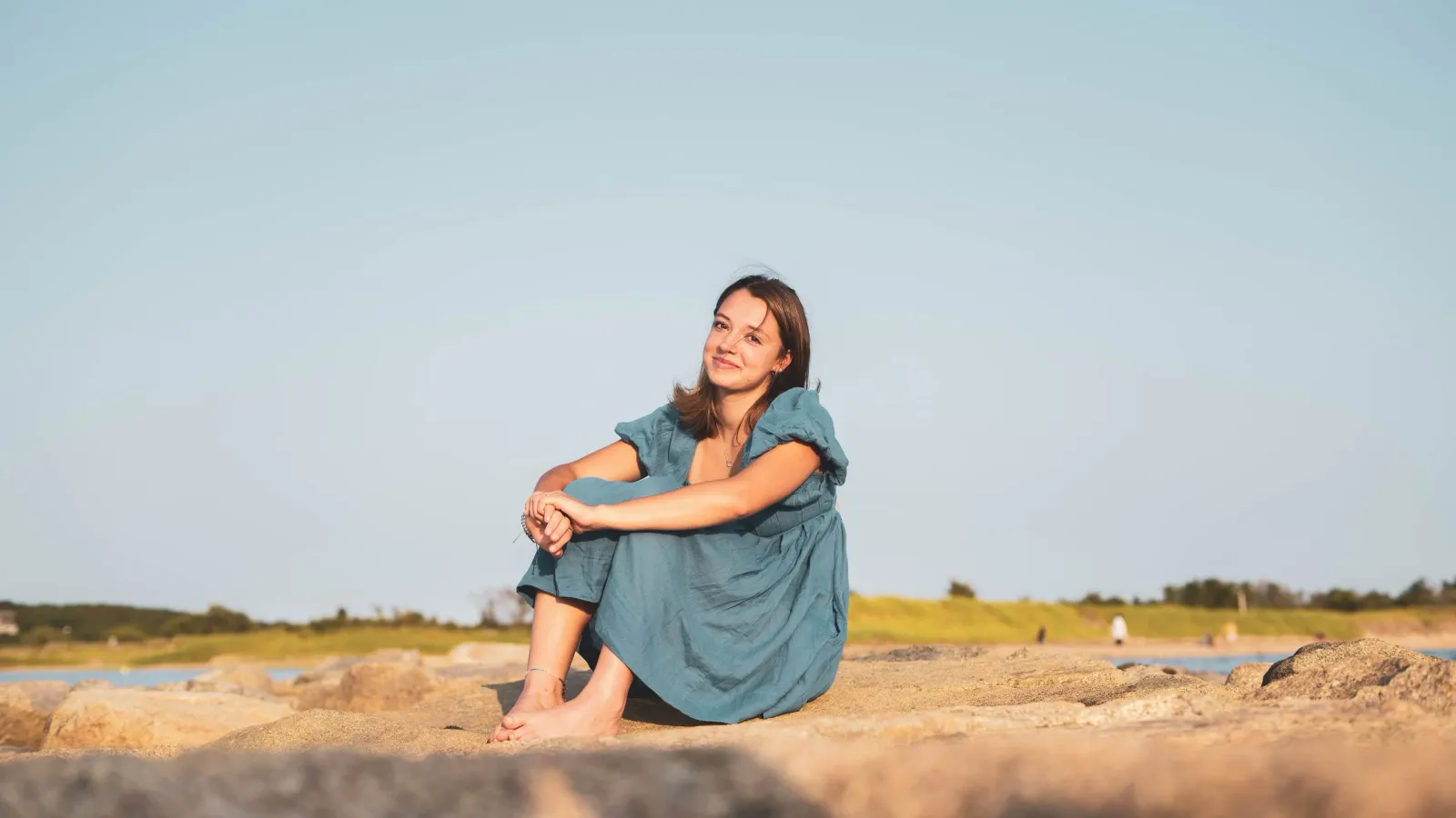 A young woman sitting on beach rocks in a teal dress, smiling at the camera with a marsh and water behind her.