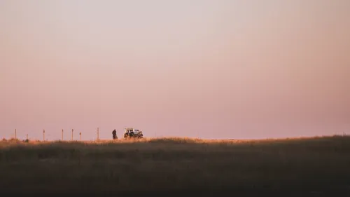 Two silhouetted figures and a parked vehicle on a grassy dune ridge at dusk.