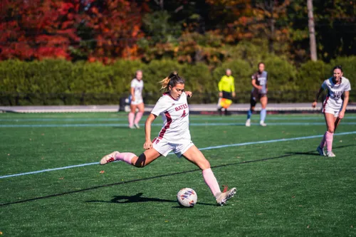 A Bates College women's soccer player striking the ball mid-kick on an autumn field lined with red foliage.