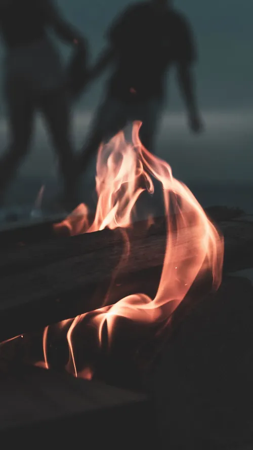 Close-up of a driftwood fire on the beach with two silhouetted figures walking past in the background.
