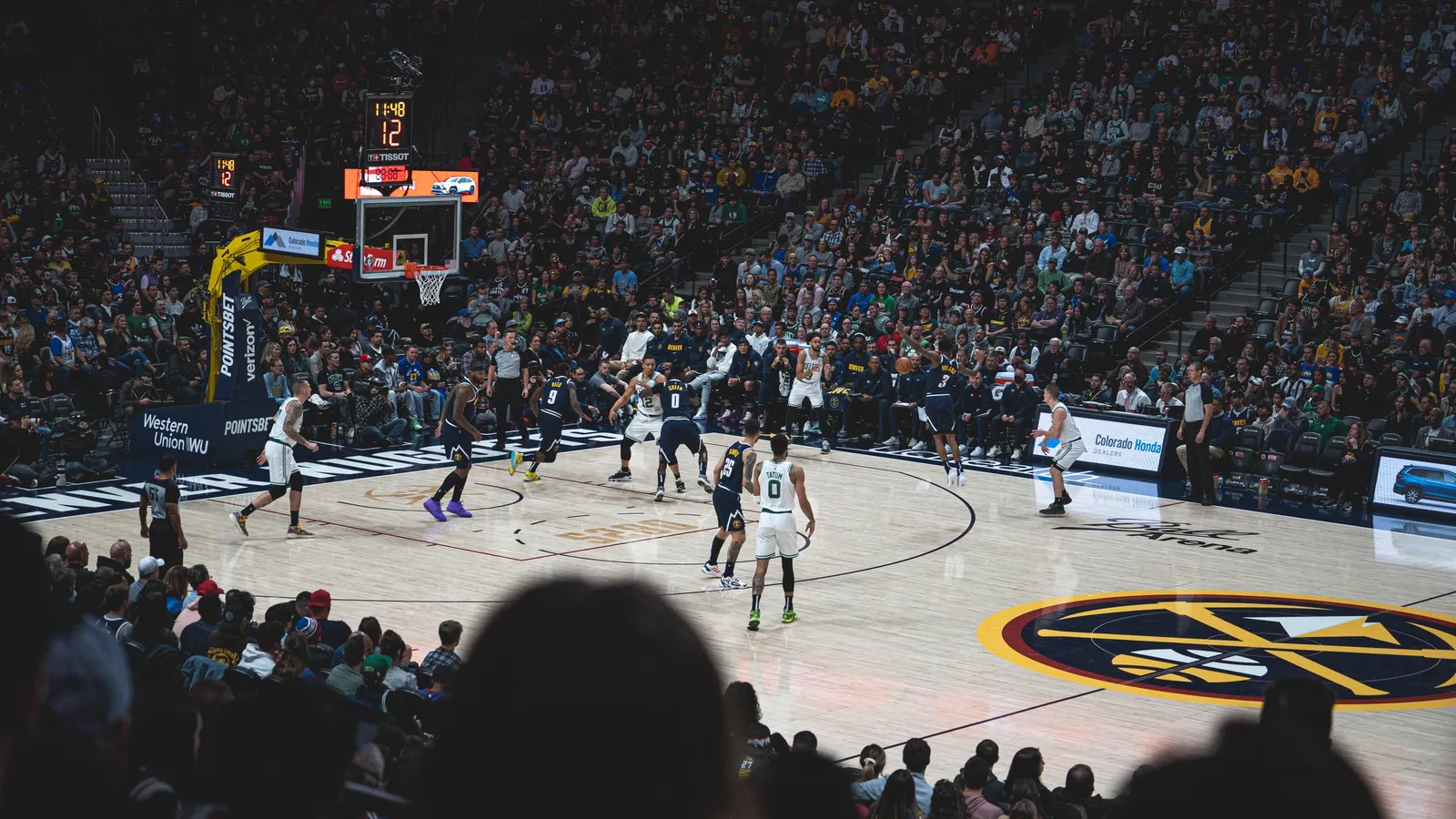 Wide view of an NBA arena during a Celtics-Nuggets game, players positioned around the top of the key in front of a packed crowd.