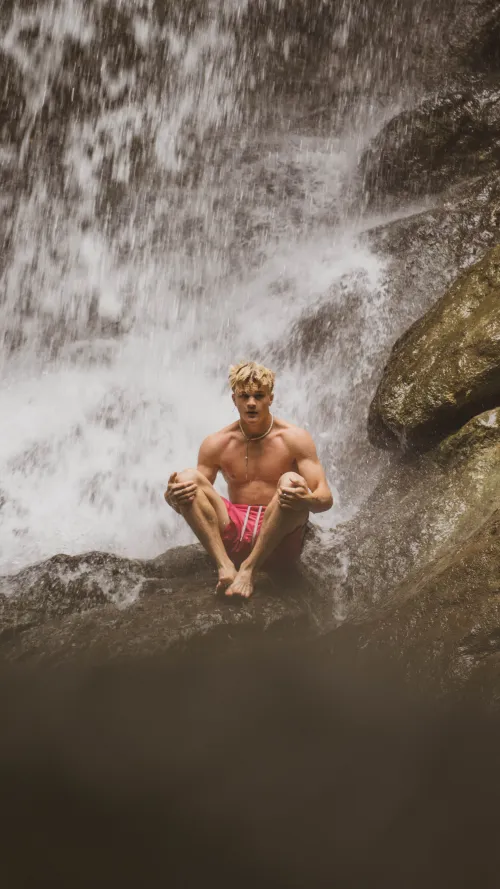 A shirtless man in pink shorts sitting on a rock beneath a waterfall, water cascading behind him.