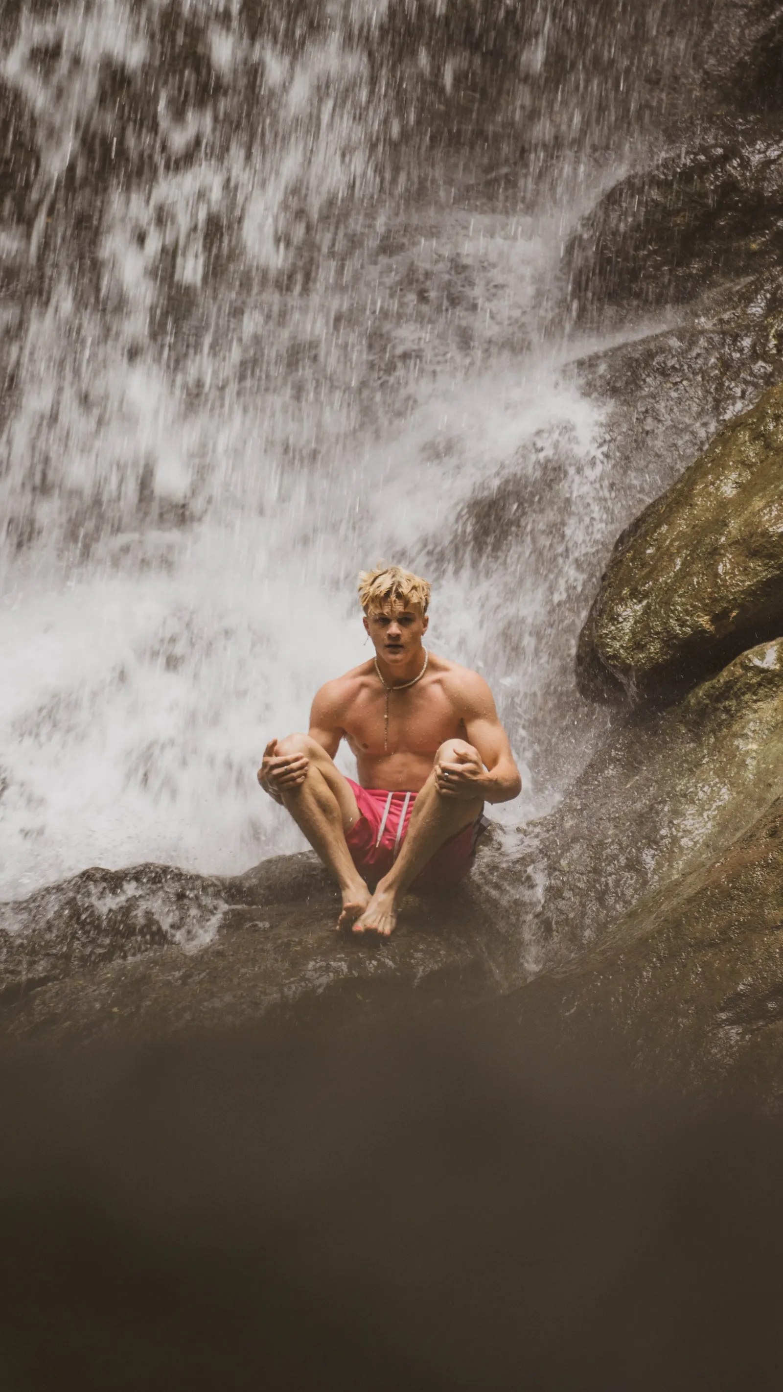 A shirtless man in pink shorts sitting on a rock beneath a waterfall, water cascading behind him.