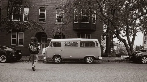 Black-and-white street scene of a vintage VW bus parked in front of a brick apartment building, a person walking past.