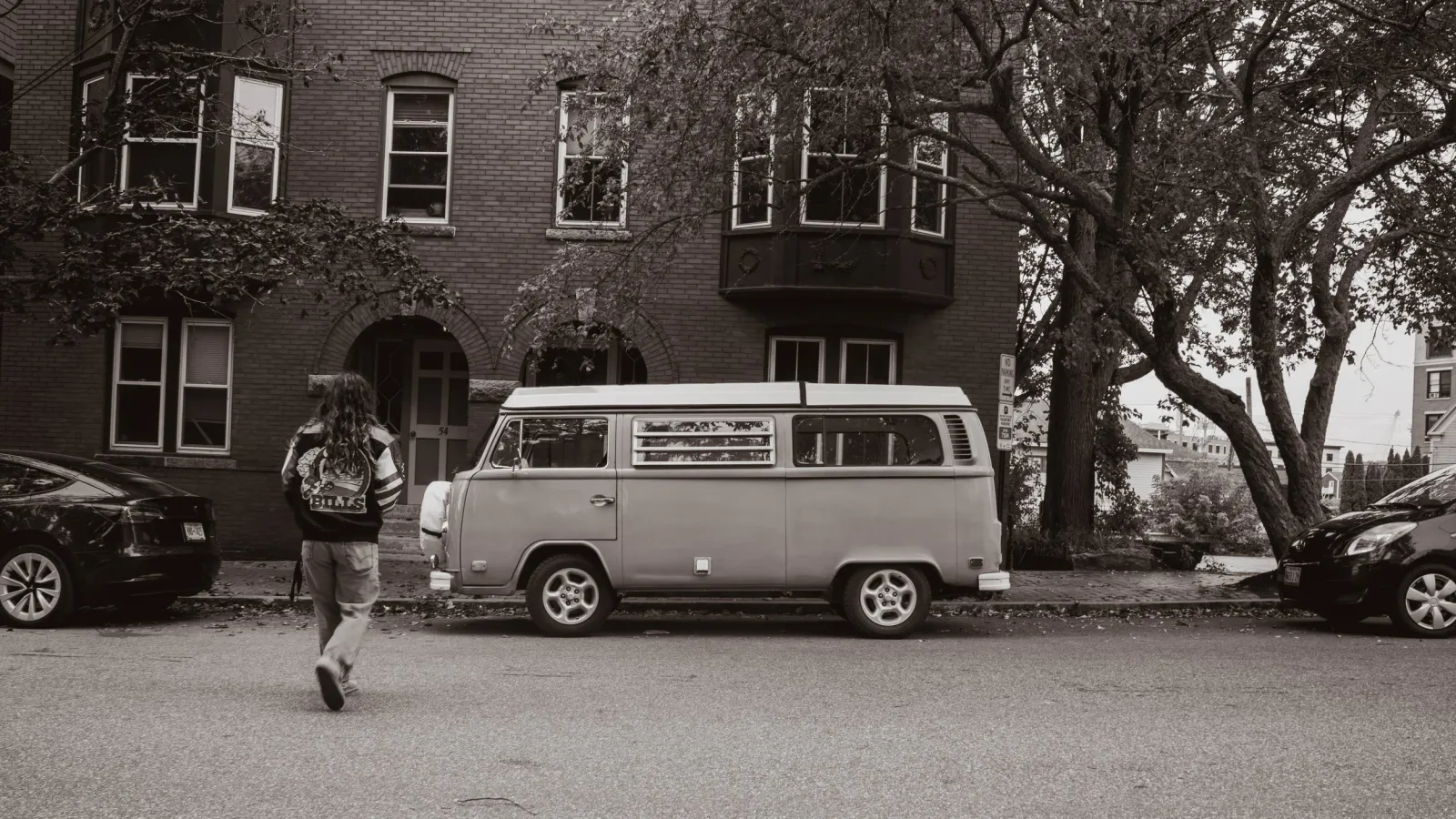 Black-and-white street scene of a vintage VW bus parked in front of a brick apartment building, a person walking past.