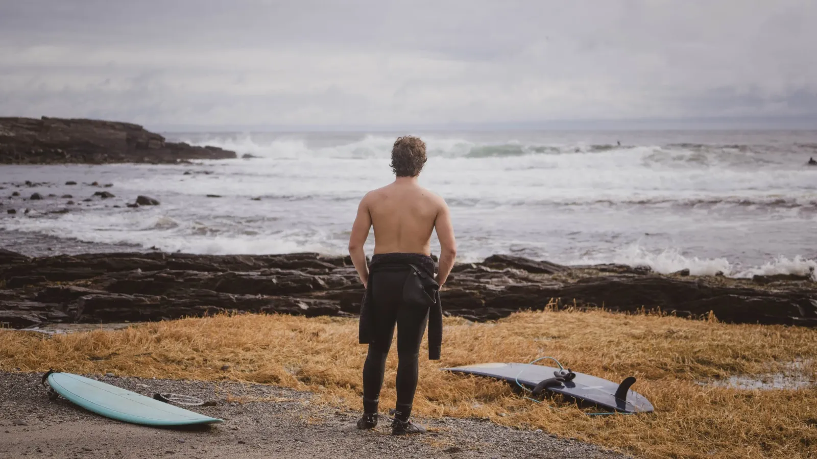 A shirtless man in a wetsuit standing on rocky shoreline grass, facing rough surf.