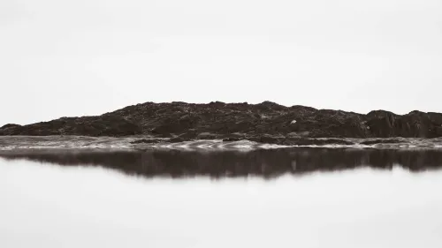 Black-and-white minimalist seascape of a rocky outcrop reflected in still water beneath a hazy, overexposed sky.