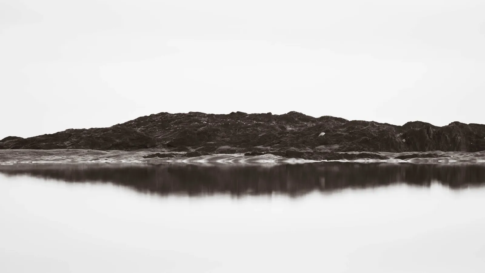 Black-and-white minimalist seascape of a rocky outcrop reflected in still water beneath a hazy, overexposed sky.