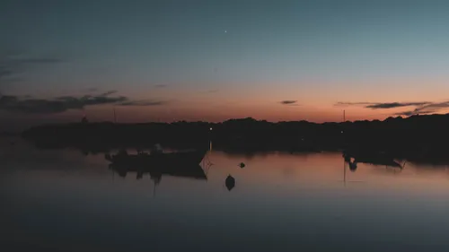 A fishing boat and buoy silhouetted on still water at dusk, shoreline and scattered lights beyond.