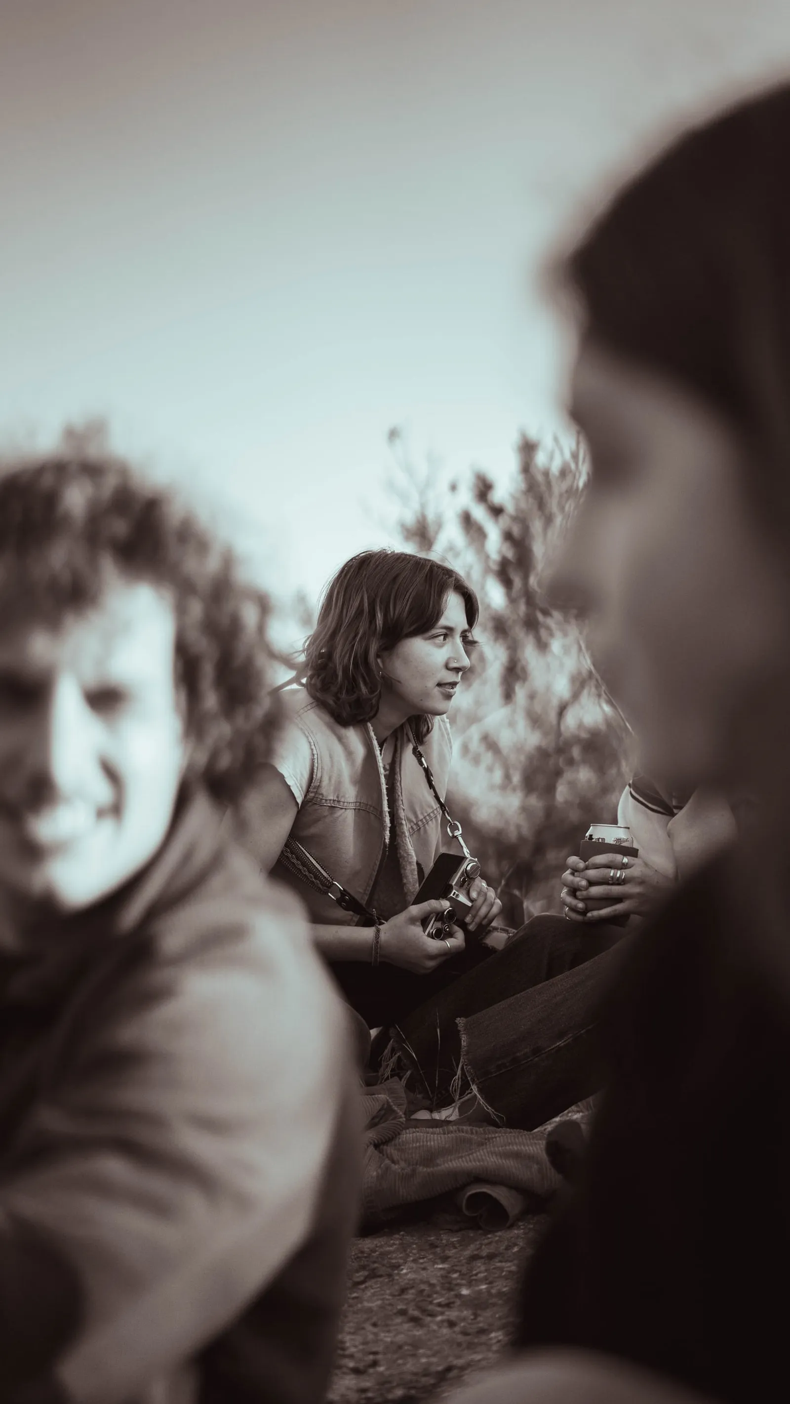 Black-and-white candid of a woman holding a film camera and a drink, sitting outdoors with friends, one in soft focus in the foreground.