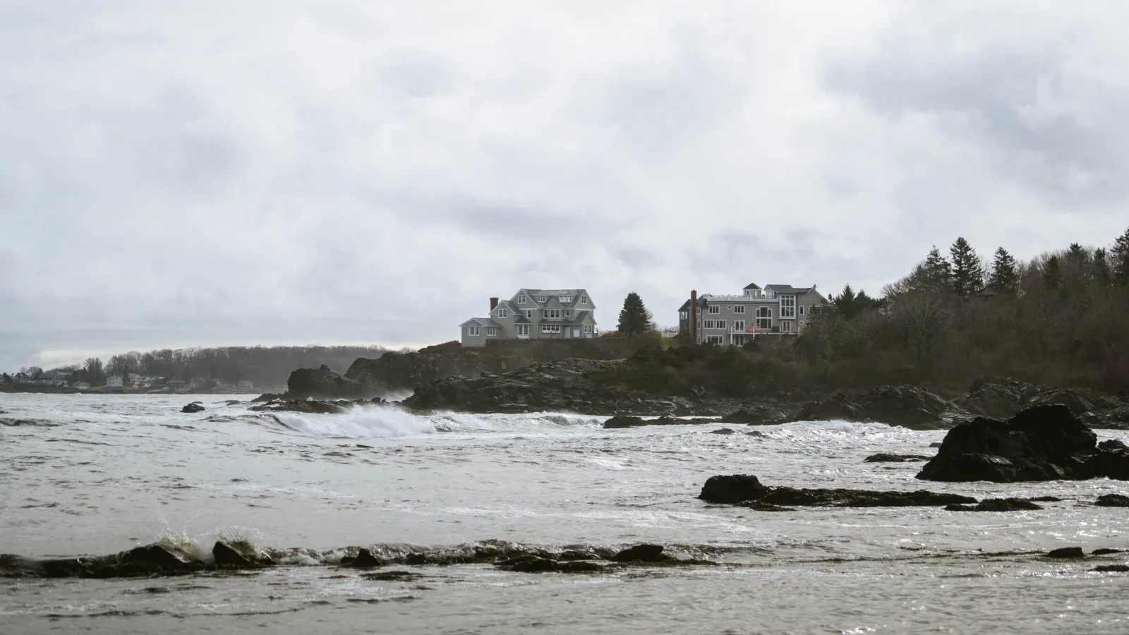 A large shingle-style coastal mansion on a rocky point, waves breaking below under an overcast sky, a coastal village visible in the distance.