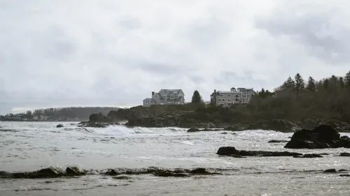 A large shingle-style coastal mansion on a rocky point, waves breaking below under an overcast sky, a coastal village visible in the distance.