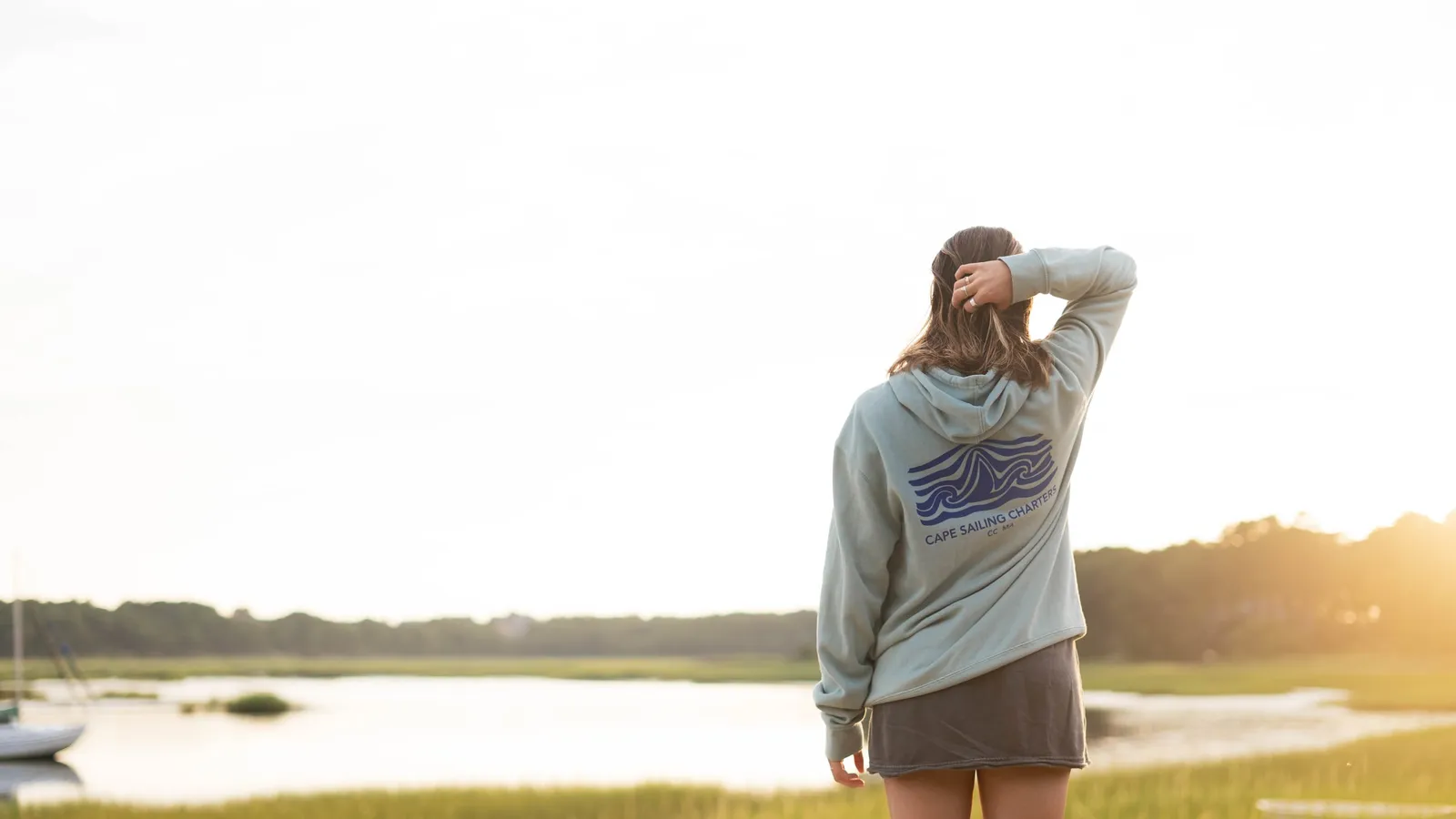 A woman seen from behind in a sailing-charter hoodie, looking out over a marsh and moored sailboats at sunset.