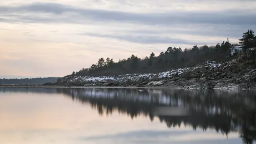 Calm winter shoreline reflecting a snow-dusted rocky bluff and evergreen treeline at dusk.