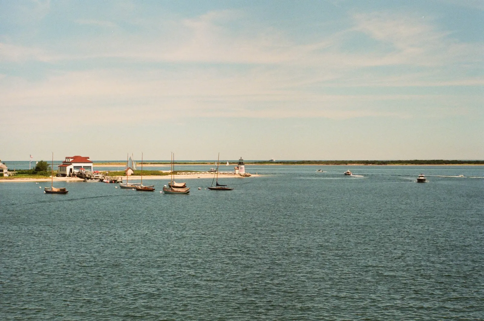 Film photo of small sailboats moored near a red-roofed boathouse and a lighthouse marking a harbor entrance.