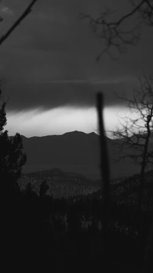 Black-and-white nighttime mountain silhouette beneath a band of storm cloud, framed by bare tree branches.