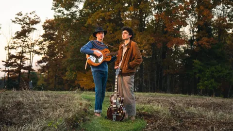 Two young men standing in an autumn field at dusk, one holding a resonator guitar and wearing a wide-brim hat, the other holding an electric guitar.