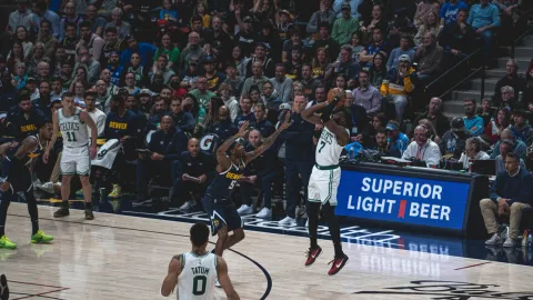 A Boston Celtics player rising for a jump shot over a Denver Nuggets defender in front of a packed arena crowd.